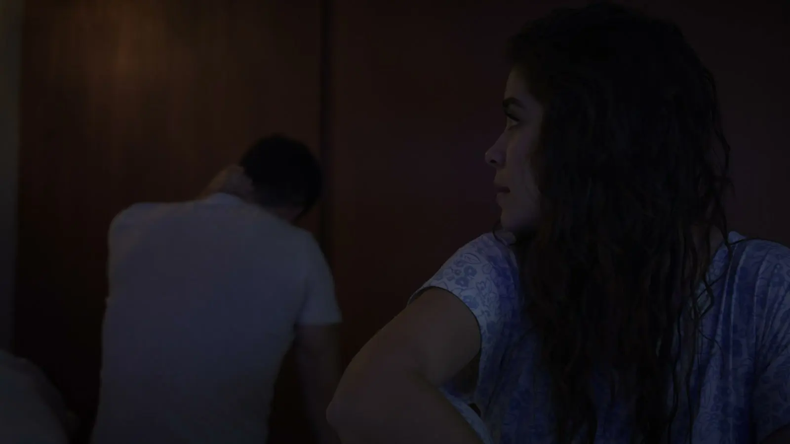 A woman looking aside while a partner sits turned away in a dark room