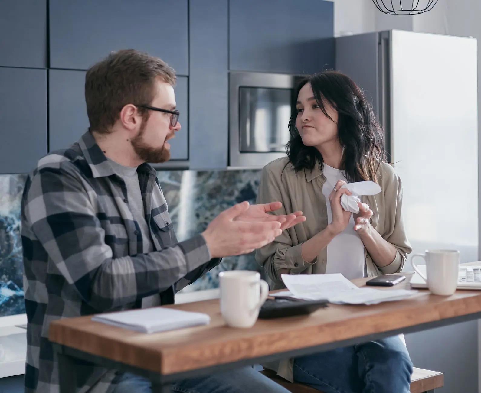 Two people talking at a kitchen table with mugs and papers between them.