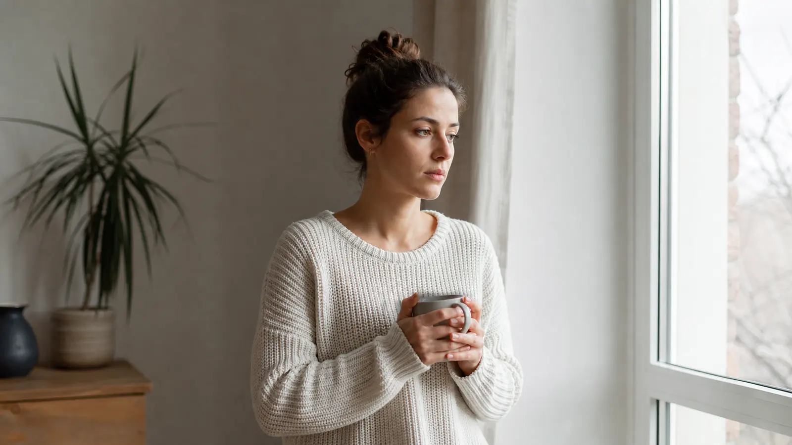 A person holding a mug and reflecting beside a bright apartment window